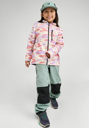 Bambino sorridente che indossa una giacca mimetica pastello, un cappellino verde menta, pantaloni verde chiaro e nero e scarpe da ginnastica blu scuro su uno sfondo semplice.