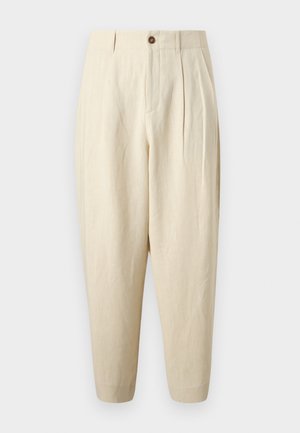 Homme portant une chemise blanche ample à manches courtes boutonnée, un pantalon beige à jambes larges et des espadrilles noires, debout avec une main dans la poche.