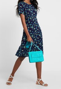 Laivansiniset vaatteet, joissa on kukkakuvio, lyhyet hihat ja liehuva hame; kantaa turkoosia teksturoitua käsilaukkua ja käyttää hopeanvärisiä sandaaleja.