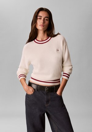 Femme portant un pull crème avec des rayures bordeaux au niveau du col, des poignets et de l'ourlet, associé à un jean foncé et une ceinture noire, mains dans les poches.
