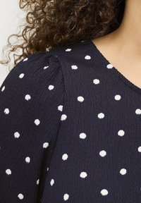 Ciemnoniebieska bluzka o teksturowej powierzchni w białe kropki, z okrągłym dekoltem i bufiastymi rękawami.