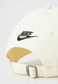 Balta beisbolo kepuraitė, pagaminta iš medvilnės, su juodu siuvinėtų "Nike" logotipu priekyje ir metaliniu reguliuojamu dirželiu gale.