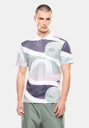 Modello maschile con capelli corti che indossa una polo con motivo astratto multicolore e pantaloncini verdi con loghi EA7, in piedi davanti a uno sfondo semplice.