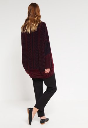 Femme aux longs cheveux portant un pull en maille ample rouge foncé et noir, un pantalon noir et des ballerines noires, debout devant un fond uni.