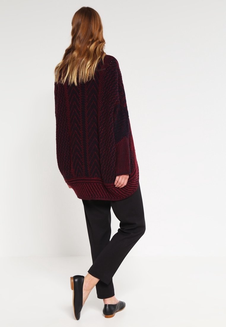 Femme aux longs cheveux portant un pull en maille ample rouge foncé et noir, un pantalon noir et des ballerines noires, debout devant un fond uni.