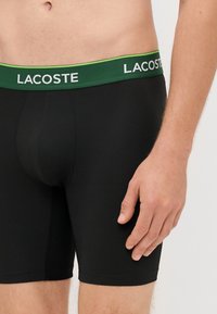 Črne bokserice z zelenim pasom, na katerem je v beli barvi logotip "LACOSTE". Gladka tekstura in oblikovanje do sredine stegen.