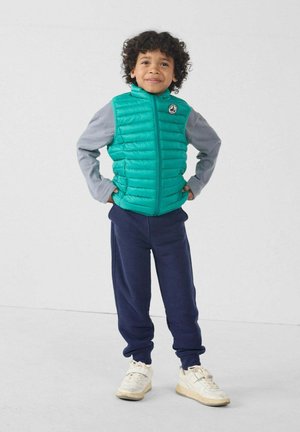 Chaleco acolchado color azul verdoso sobre camisa gris de manga larga, combinado con pantalones deportivos azul marino y zapatillas blancas. El chaleco tiene costuras verticales y un logo.