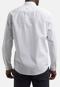 Homme portant une chemise blanche à manches longues et un pantalon foncé, vu de dos sur un fond uni.