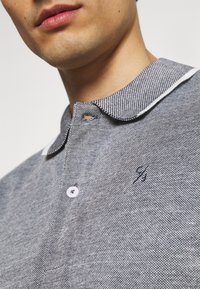 Grå poloskjorte med et strukturert mønster, hvit krage og en liten blå logo på brystet. Har en knappestolpe med to hvite knapper.