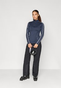 Námořnická mikina na zip s dlouhým rukávem a bílými bočními pruhy, ve spojení s černými kalhotami a lyžařskými botami. Oblečení zahrnuje lyžařskou čepici drženou v rukou.