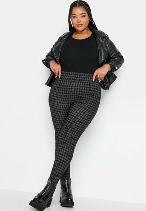 Schwarze Kunstlederjacke, schwarzes enges Oberteil und Leggings im Hahnentrittmuster in Grautönen. Klobige schwarze Stiefeletten mit Reißverschlüssen.