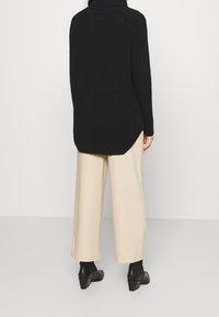 Person, die von hinten zu sehen ist und einen schwarzen strukturierten Pullover, beige weite Hosen und schwarze knöchelhohe Stiefel mit Absatz trägt.