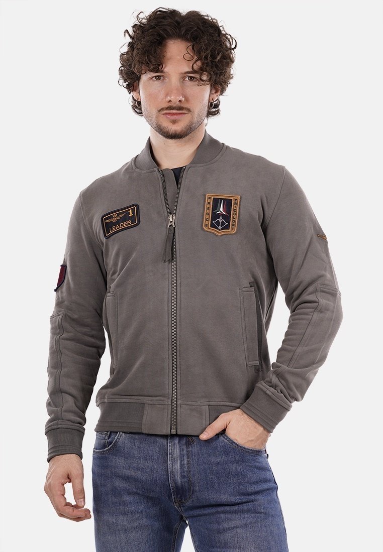 Giacca bomber grigia con zip frontale, polsini a costine e tasche laterali. Caratterizzata da toppe ricamate sul petto e sulla manica per dettagli decorativi.