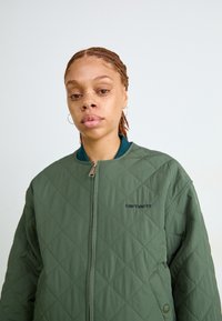 Maslinasto zeleni krečasti kaput s patentnim zatvaračem, s okruglim izrezom i crnim Carhartt logotipom na lijevoj strani prsnog područja.