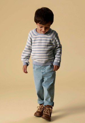 Jeune garçon portant un pull rayé bleu et blanc, un jean bleu clair et des bottes marron, regardant vers le bas sur un fond beige.