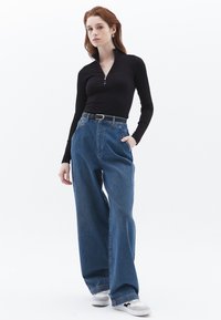Zwarte lange mouwen top met een halflange rits kraag, gecombineerd met hoge taille, wijde blauwe spijkerbroeken. De outfit bevat een zwarte riem en witte sneakers.