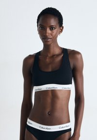 Svart sports-bh og matchende truse med et bredt hvitt langt belte med gjentatt "Calvin Klein"-logo. Glatt, elastisk stoff, tettsittende design.