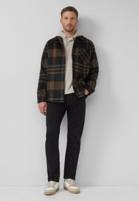 Kostka flanelowa w kolorze czarnym, brązowym i beżowym, narzucona na jasnoszary hoodie, w połączeniu z ciemnymi spodniami i kremowymi trampkami.