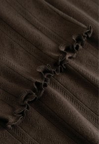 Hnedá pletená tkanina s volánovým okrajom a jemným vzorom diamantov a čiar, ktorý zvýrazňuje textúrovaný, útulný vzhľad.