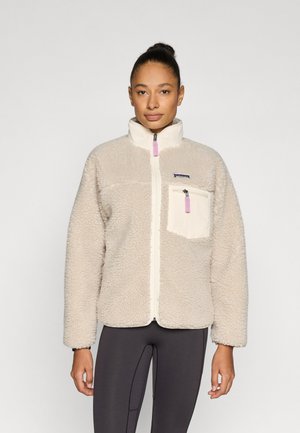 Beige fleecejakke med høy krage, frontglidelås og et lomme med lapp. Har en kontrasterende kremfarget lomme og rosa glidelåstrekk.