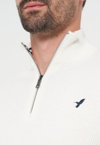 Gros plan sur le bas du visage et le cou d’un homme portant un pull blanc côtelé avec une fermeture éclair courte et un petit logo oiseau noir sur la poitrine.