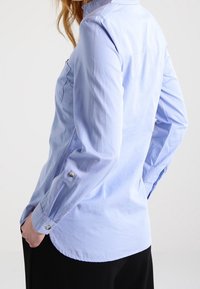 Ljusblå skjorta med långärmar, som har en subtil lyster, krage och dekorativa knappdetaljer på manschetterna.
