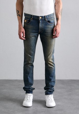 Dark blue denim jeans with a relaxed fit, featuring light fading, front pockets, and gold hardware. Paired with white sneakers.