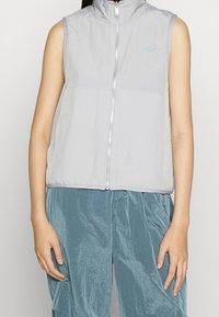 Ljust grå ärmlös väst med hög krage, dragkedja framtill och subtil logotypdetalj, tillverkad av ett texturerat, lätt material.