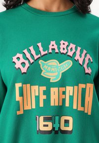 Felpa verde in cotone con testo stampato audace: "BILLABONG," "Surf Africa," e "1640" in rosa e arancione con accenti blu.