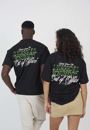 Dwie osoby stoją tyłem, ubrane w czarne koszulki z zielonym i białym napisem: "IL VIAGGIO È IL MONUMENTO Out of Office."