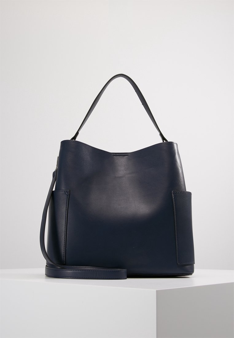 Sac à main en cuir bleu marine foncé avec une poignée supérieure unique et une bandoulière amovible, placé sur un piédestal blanc sur un fond uni.