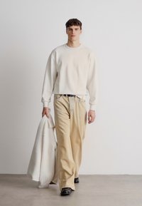 Κρέμα φούτερ με χαλαρή εφαρμογή, συνδυασμένο με ανοιχτό μπεζ παντελόνια. Το μοντέλο κρατάει ένα φουντωτό λευκό παλτό, φορώντας μαύρα παπούτσια. Μινιμαλιστικός σχεδιασμός.