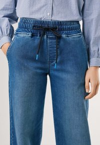 Denim hlače za trening imaju opušteni kroj, plavu boju, elastičnu struk s vezicom i bočne džepove. Tekstura tkanine je glatka.