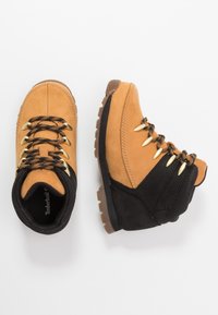 Botas de senderismo en tono marrón y negro con cordones negros y naranjas, suelas gruesas y cuellos acolchados, mostradas desde vistas superior y lateral sobre fondo blanco.