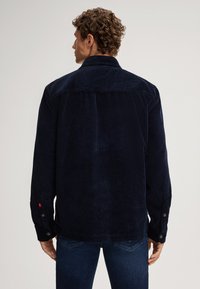 Camicia in corduroy blu navy con colletto button-down, linee verticali visibili e dettagli dei polsini abbottonati. Tessuto liscio e vestibilità rilassata.