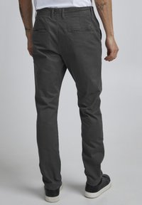 Grå bomullsbukser med slim fit, baklommer med klaffer og en glatt tekstur. Brukt med svarte sneakers mot en nøytral bakgrunn.