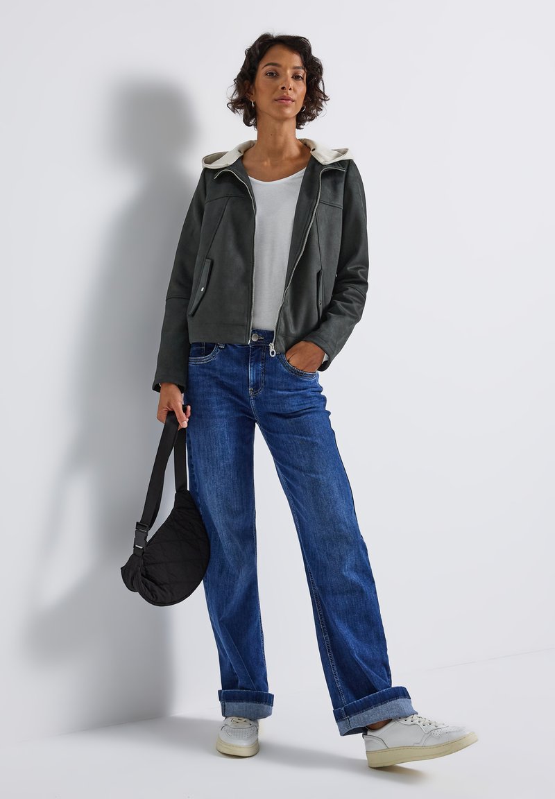 Vrouw in een donkergrijze jas over een wit shirt, blauwe omgeslagen jeans, witte sneakers, met een zwart gewatteerd tasje, staand tegen een witte muur.