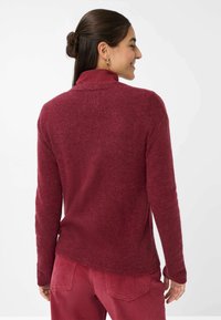 Bordeaux-farbenes Strickoberteil mit Rundhalsausschnitt und langen Ärmeln, das über eine weiche Textur und einen leicht abgerundeten Saum auf der Rückseite verfügt.