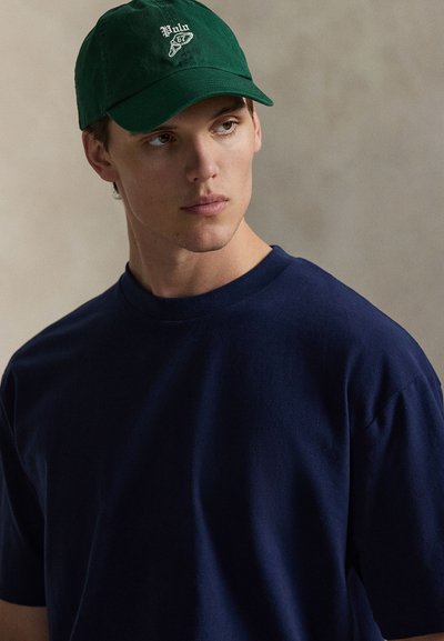 Μαύρο-μπλε βαμβακερό μπλουζάκι με κοντά μανίκια, σε συνδυασμό με σκούρο πράσινο καπέλο μπέιζμπολ με λευκό κεντημένο το κείμενο "Polo" στο μπροστινό μέρος.