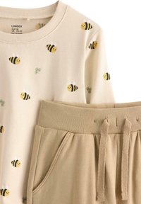 Beige sweatshirt med gule bi mønstre og grønne detaljer, parret med lysebrune bukser med ribbet talje og snor.