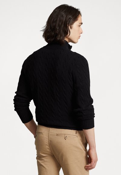 Black cable-knit sweater with a high collar, ribbed cuffs, and a fitted design, paired with light khaki pants. Textured fabric detail visible.