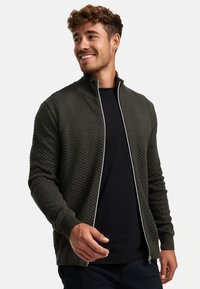 Rits-cardigan in donkergroen met een zigzagpatroon, met een hoge kraag en een contrasterende rits. Draag het over een zwart shirt.