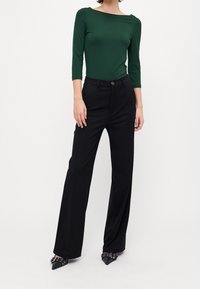 Groene lange mouw top gecombineerd met hoge taille zwarte wijde broek. Broek heeft een knoopsluiting; schoenen zijn zwart met een puntige neus.