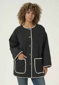 Svart yllekappa med vit scallopstickning, oversized passform, framfickor och stora svarta knappar. Strukturerad tyg med avslappnad design.