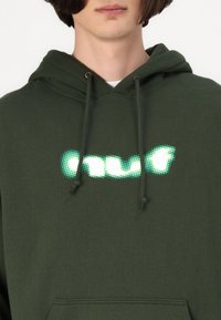 Hanorac verde închis, cu un logo mare din text alb strălucitor, accentuat cu puncte. Are șnururi și o buzunar frontal. Material dintr-un amestec de bumbac.