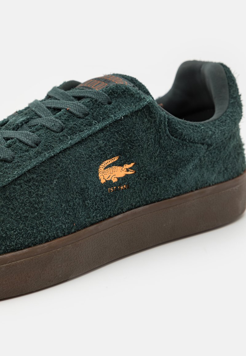 Zielone zamszowe sneakersy z brązową gumową podeszwą, z płaskimi sznurówkami i wytłoczonym pomarańczowym logo krokodyla z boku.