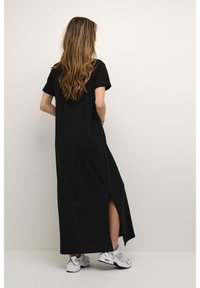 Robe longue noire à manches courtes avec fente latérale, confectionnée dans un tissu doux. Associée à des baskets blanches de sport, vue de dos.