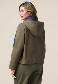 Giacca verde oliva con cappuccio, caratterizzata da dettagli in denim blu sulle spalle, tessuto testurizzato e vestibilità rilassata in un design corto.