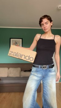 Cutie de carton cu "zalando" imprimate în litere îngroșate de culoare gri. Modelul poartă un top negru fără mâneci și blugi largi de culoare bleu deschis, având un centură neagră.