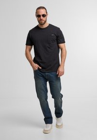 Czarna koszulka z krótkim rękawem z kieszonką na piersi, w połączeniu z niebieskimi dżinsami i jasnymi sneakersami. Model nosi okulary przeciwsłoneczne, stoi w pozycji wyprostowanej.